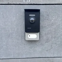 Zamontowany, czarny wideodomofon Philips z kamerą i przyciskiem dzwonka na szarym, betonowym murze. Widoczny refleks osoby w obiektywie kamery.