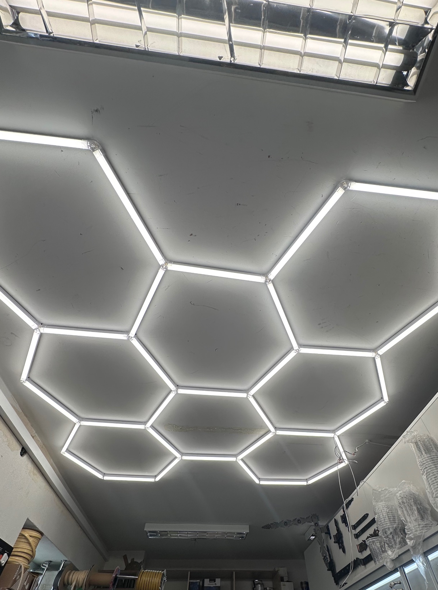 Sufit z heksagonalnym oświetleniem LED, widok z dołu. Białe światło tworzy geometryczny wzór na szarym tle, oświetlając wnętrze pomieszczenia.