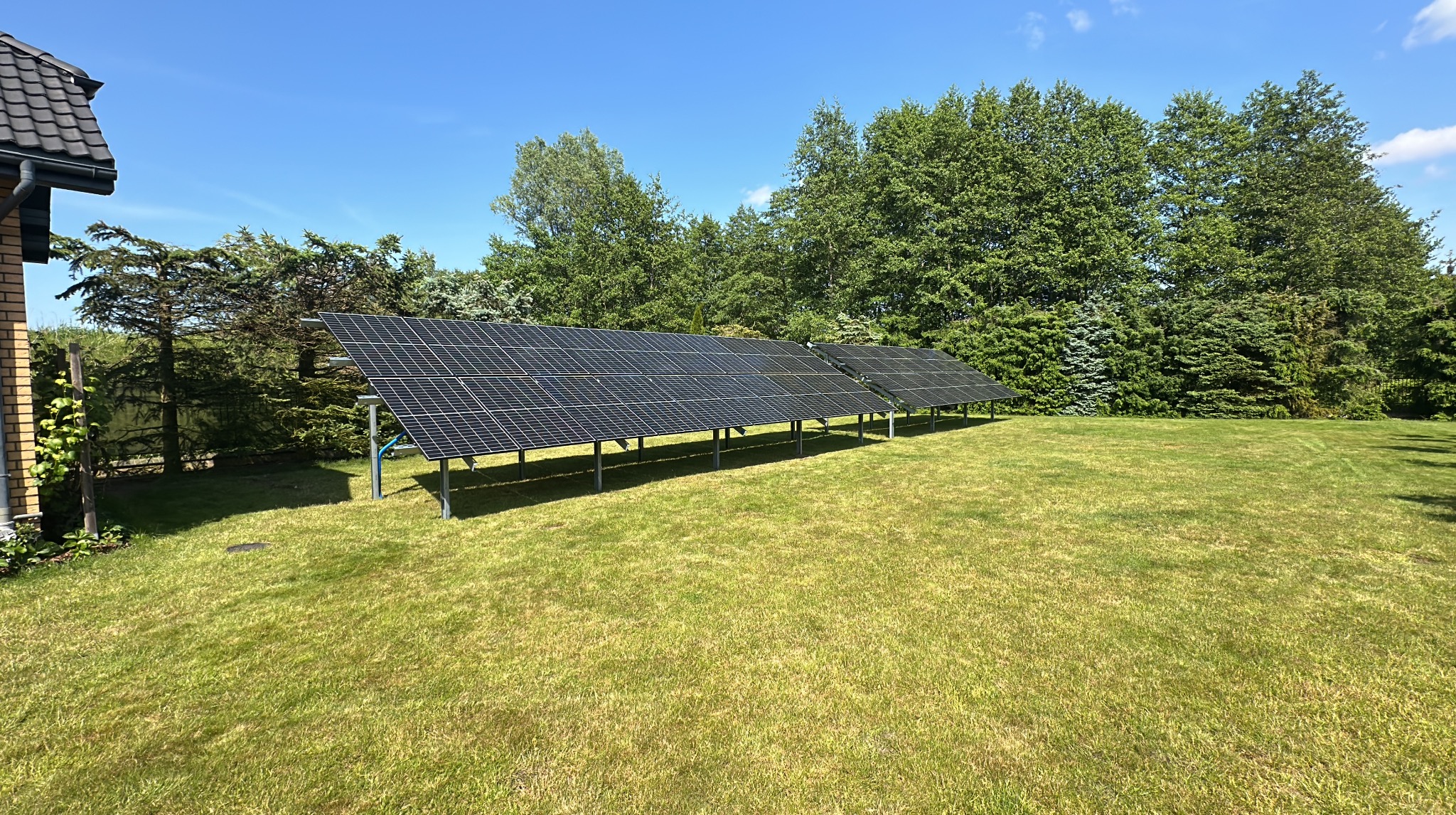 Instalacja paneli słonecznych na trawniku przed domem, widok z perspektywy trawy, słoneczny dzień, wysokie drzewa w tle, czyste niebo.