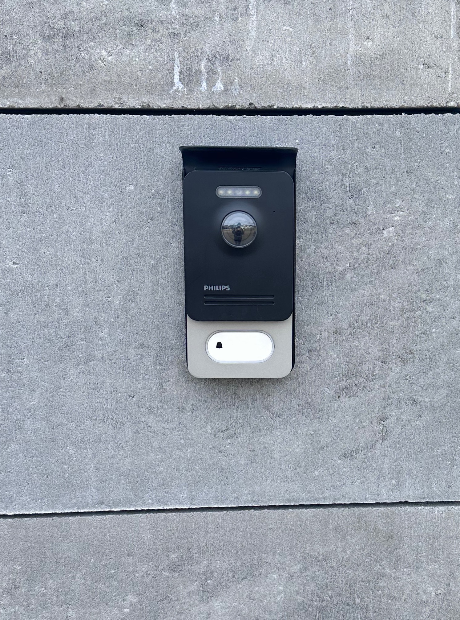 Zamontowany, czarny wideodomofon Philips z kamerą i przyciskiem dzwonka na szarym, betonowym murze. Widoczny refleks osoby w obiektywie kamery.