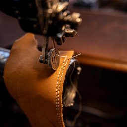 Madeinhands.pl Zenon Stokłosa - Szczegółowe ujęcie maszyny szwalniczej zszywającej brązową skórę, widoczne białe szwy. Całość w ciepłej tonacji, z ciemnym tłem.