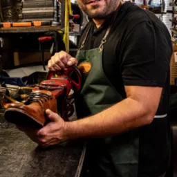 Rzemieślnik w zielonym fartuchu naprawia brązowy skórzany but przy użyciu starej, czerwonej maszyny do szewstwa w warsztacie.