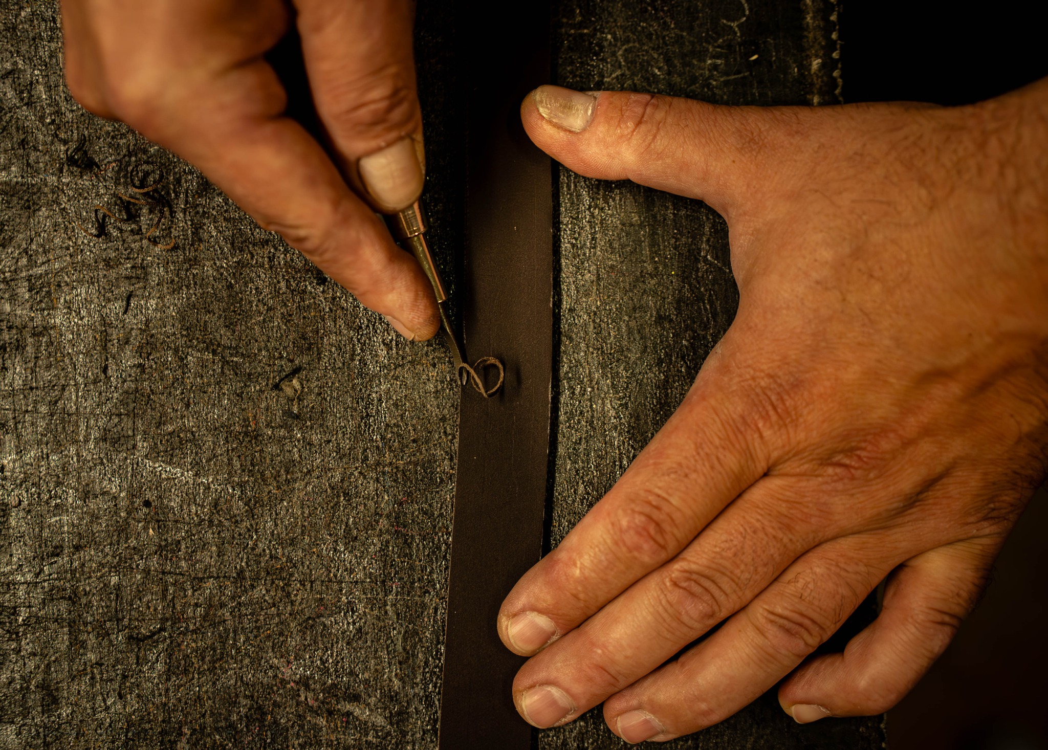 Ręczna praca: narzędzie do obróbki skóry w dłoni, precyzyjne wycinanie wzoru na pasku ze skóry na tle szorstkiego blatu. Detal rzemiosła.