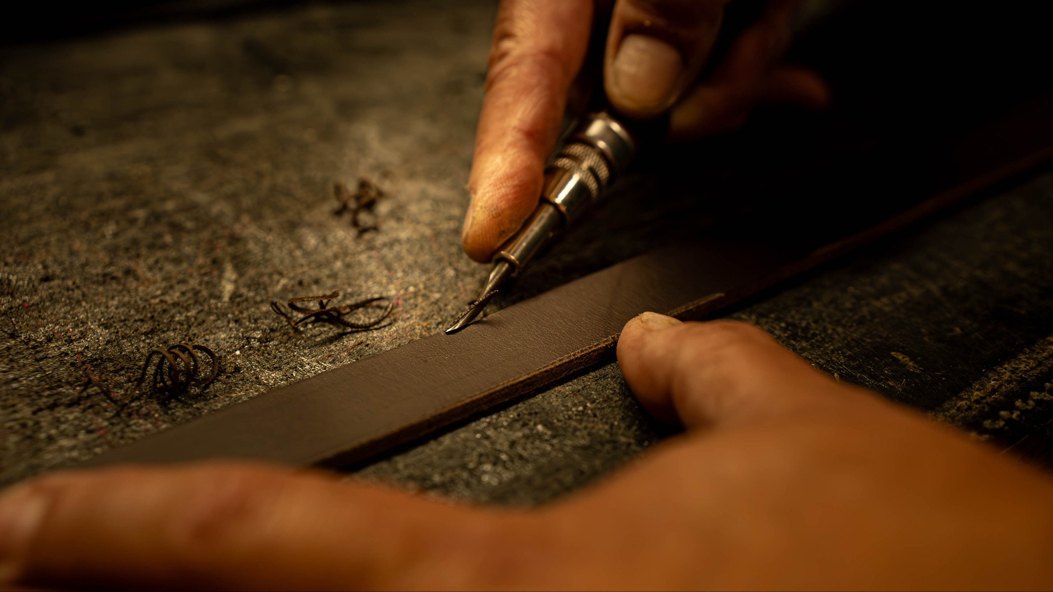 Ręczne wycinanie wzoru w brązowym pasku skórzanym za pomocą precyzyjnego narzędzia na szorstkiej powierzchni roboczej z widocznymi wiórami skóry.