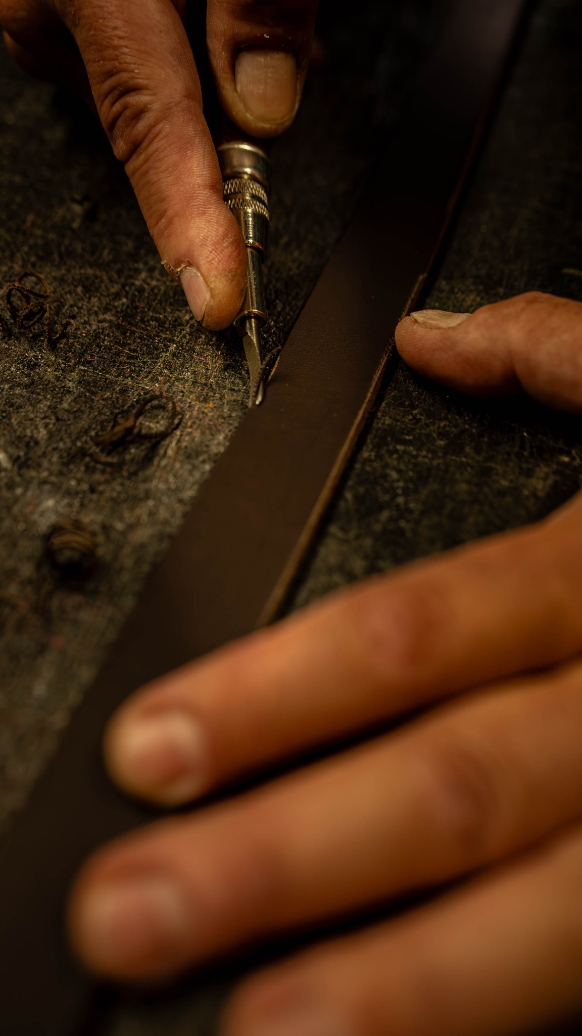 Detaliczne ujęcie dłoni precyzyjnie krojącej skórzany pasek specjalistycznym nożem na szorstkiej powierzchni roboczej. Widoczne wióry skóry.