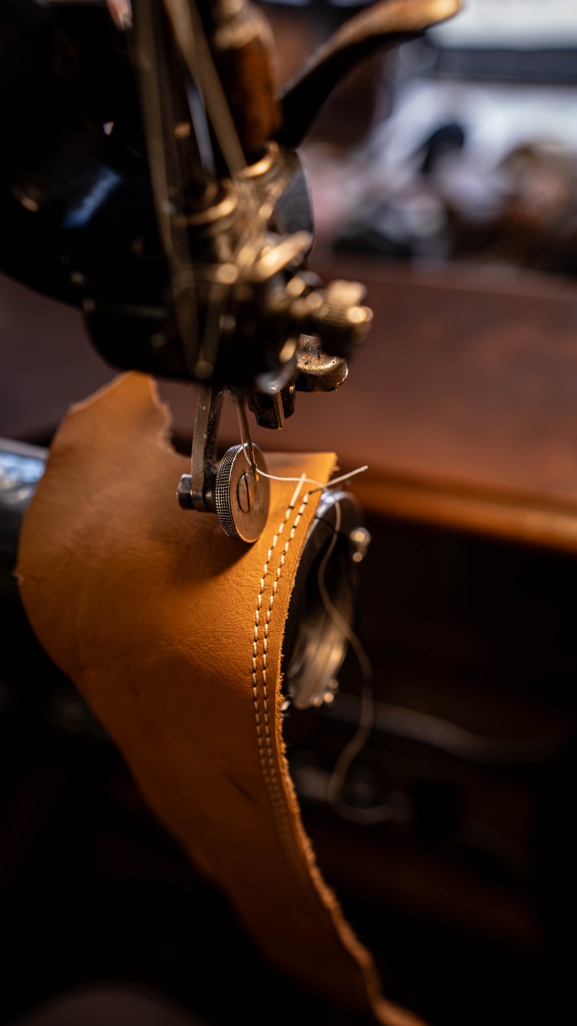 Szczegółowe ujęcie maszyny szwalniczej zszywającej brązową skórę, widoczne białe szwy. Całość w ciepłej tonacji, z ciemnym tłem.