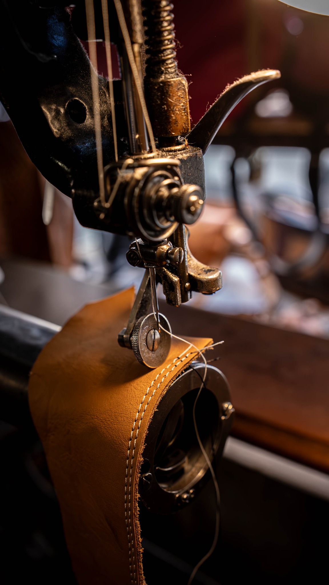 Detal maszyny szewskiej w trakcie szycia brązowej skóry, widoczne białe szwy. Oświetlenie podkreśla detale mechanizmu i fakturę materiału.