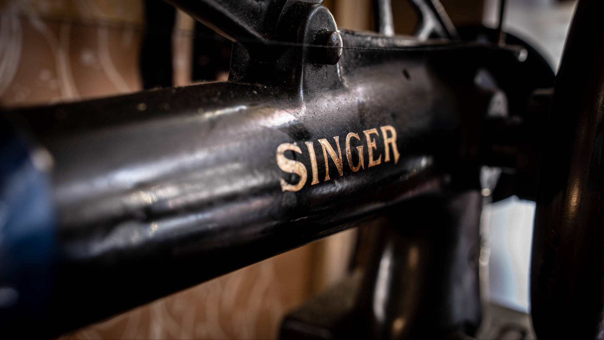 Fragment czarnej maszyny do szycia marki Singer z widocznym, złotym napisem. Ujęcie z bliska, w tle fragment drewnianej ściany.