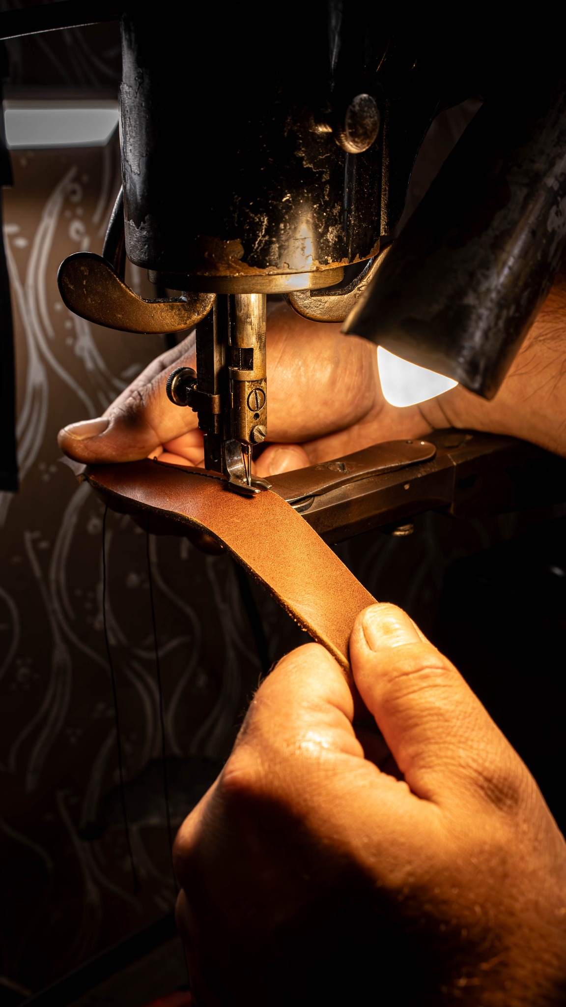 Brązowy pasek skórzany szyty na starej maszynie. Dłonie trzymają skórę pod igłą. Oświetlenie punktowe podkreśla detal. Widoczne ślady użytkowania na maszynie.