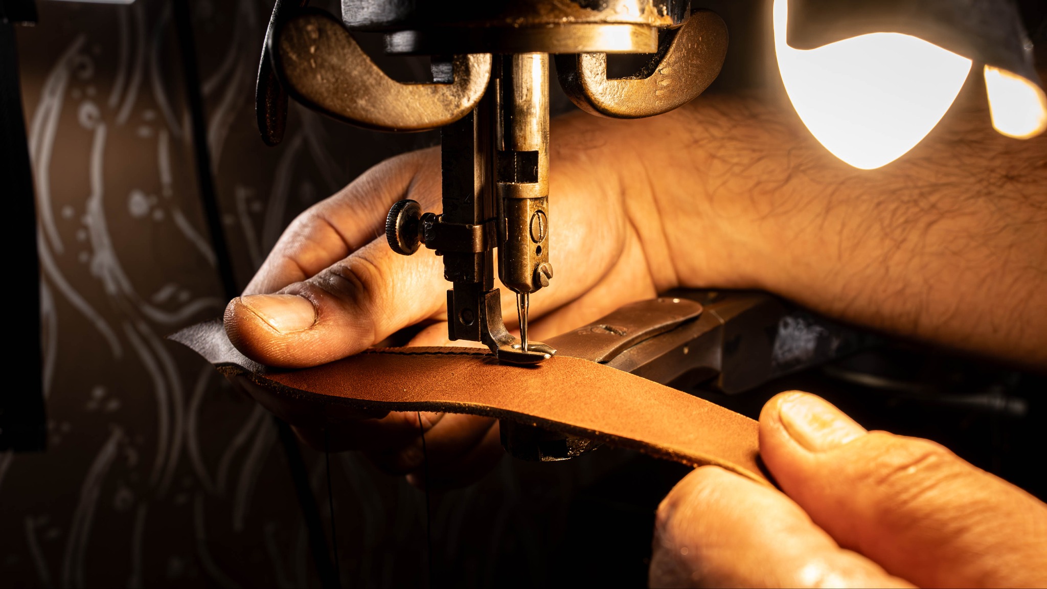 Szycie skórzanego paska na zabytkowej maszynie. Dłonie rzemieślnika prowadzą skórę pod igłą, oświetloną ciepłym światłem. Detal pracy kaletniczej.