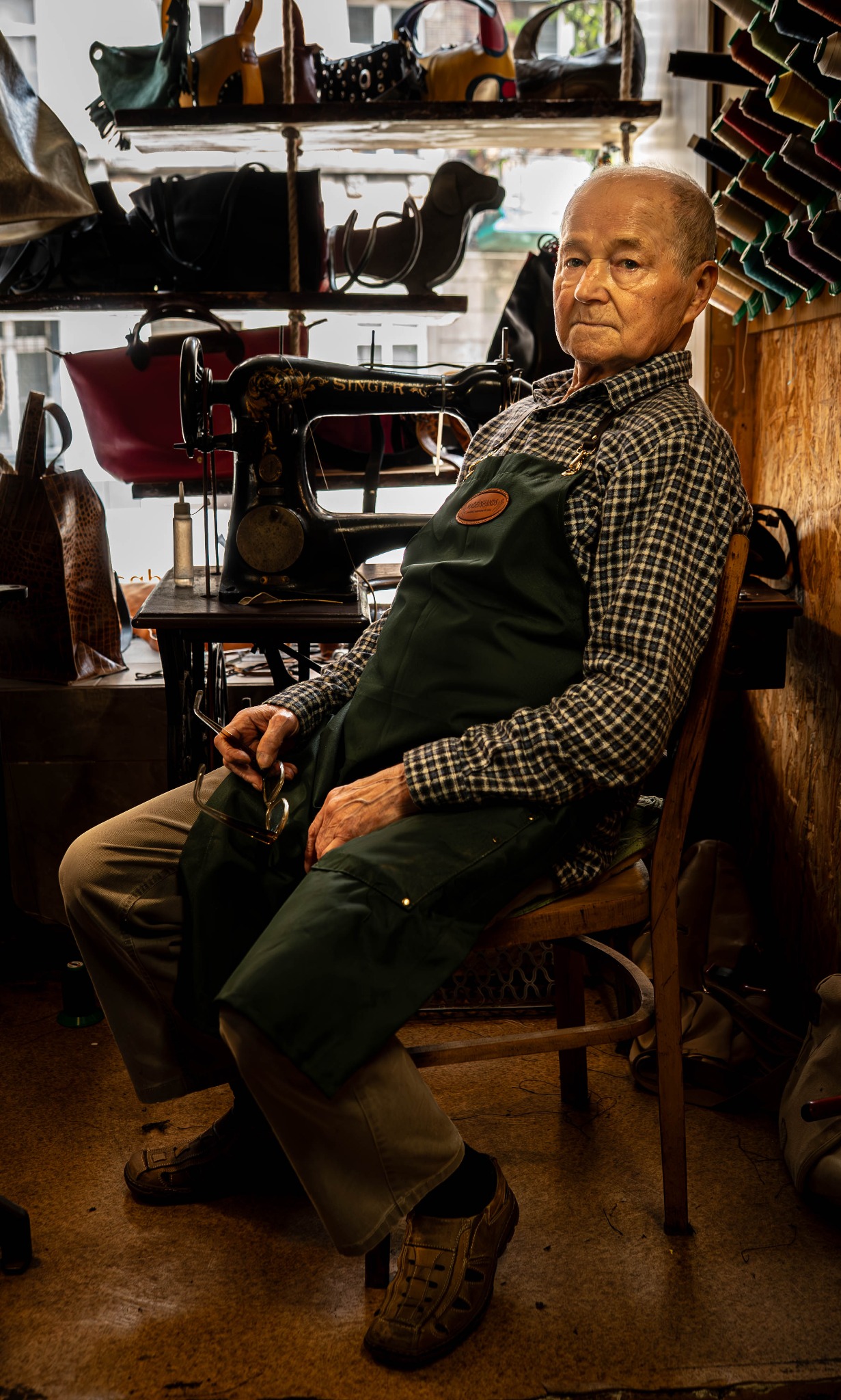 Portret kaletnika w warsztacie z maszyną Singer, w tle półki z wyrobami skórzanymi i nici. Mężczyzna w kraciastej koszuli i zielonym fartuchu siedzi na drewnianym krześle.