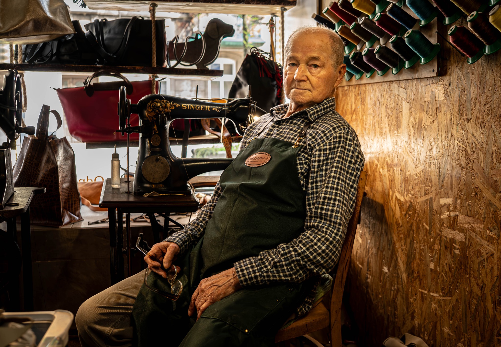 Kaletnik w warsztacie, siedzi obok starej maszyny Singer, w tle torby skórzane i nici. Portret rzemieślnika w kameralnym otoczeniu, z widocznymi narzędziami pracy.