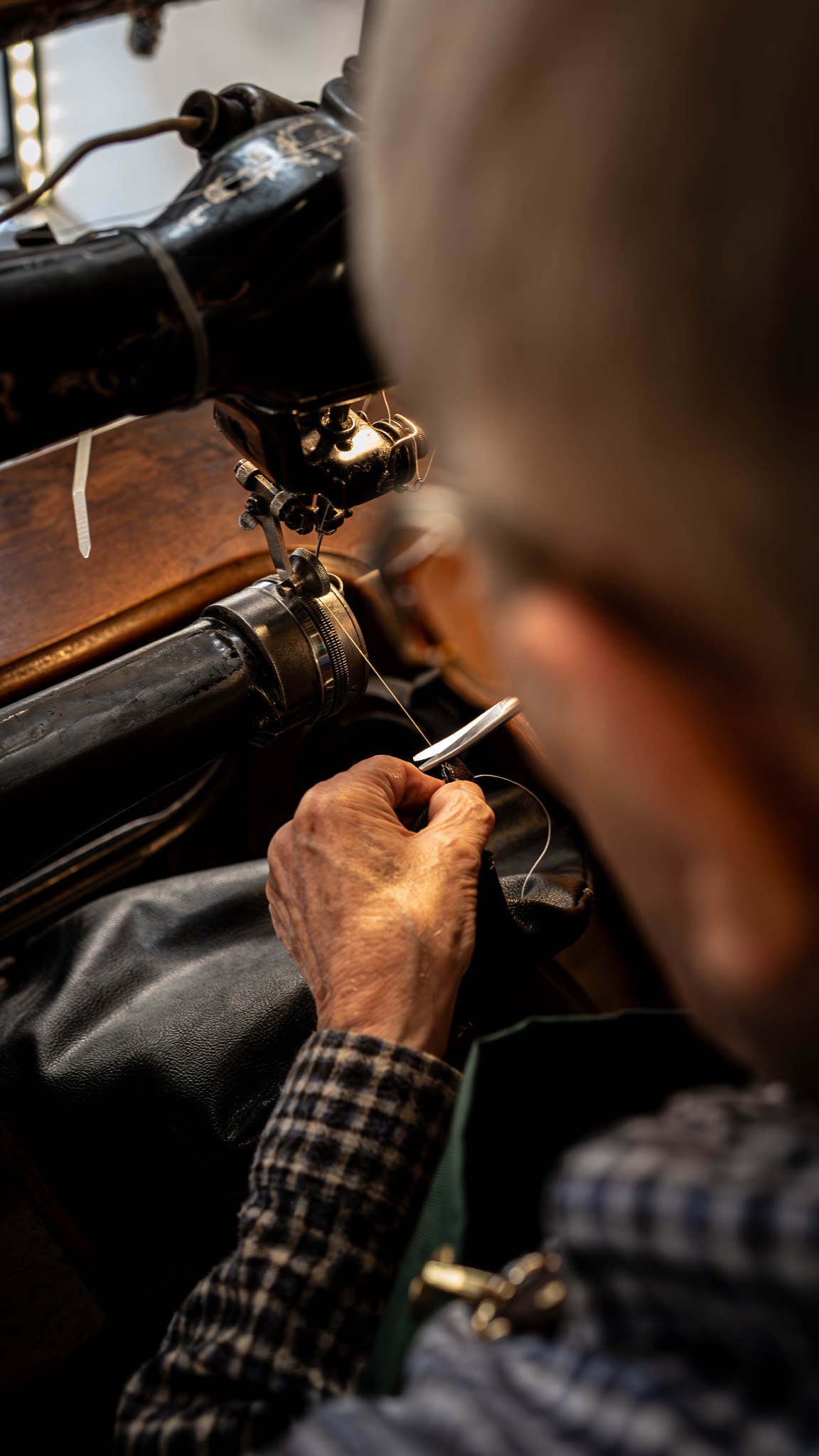 Ręka mężczyzny prowadzi skórę pod igłą starej maszyny do szycia, oświetlonej punktowo. Detal pracy rzemieślniczej w warsztacie.