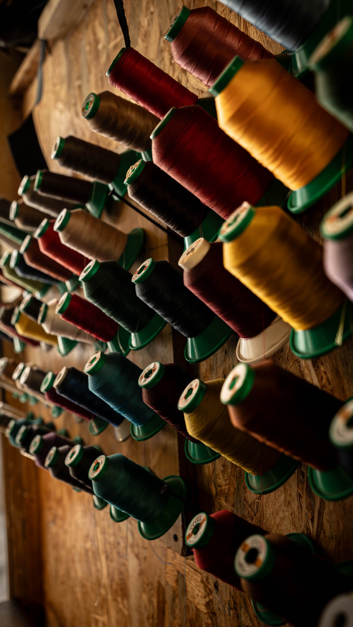 Rzędy kolorowych nici na szpulach, zawieszone na drewnianej ścianie. Nici w odcieniach czerwieni, brązu, niebieskiego i żółtego. Ujęcie z dołu, oświetlenie naturalne.