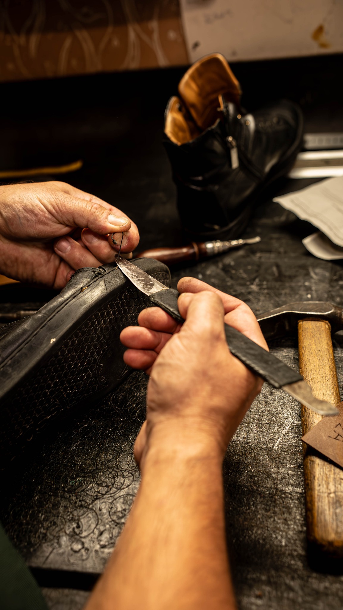 Ręczne szycie czarnego skórzanego buta przez rzemieślnika przy użyciu igły i noża na warsztacie, widoczne narzędzia kaletnicze.