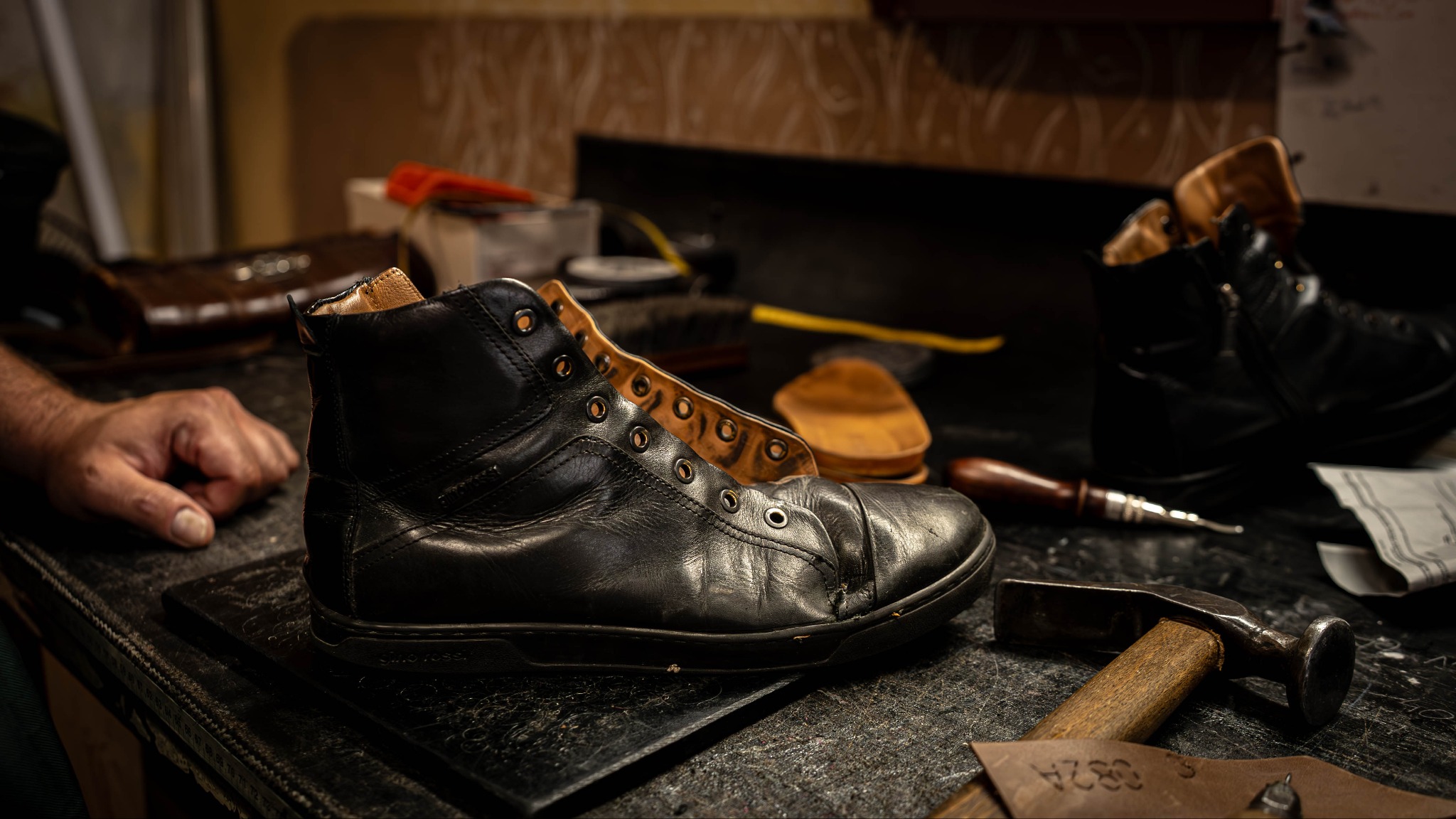 Warsztat: czarny, skórzany but na stole obok narzędzi kaletniczych i dłoni rzemieślnika. Detaliczna praca, ciemne oświetlenie, unikalny charakter.
