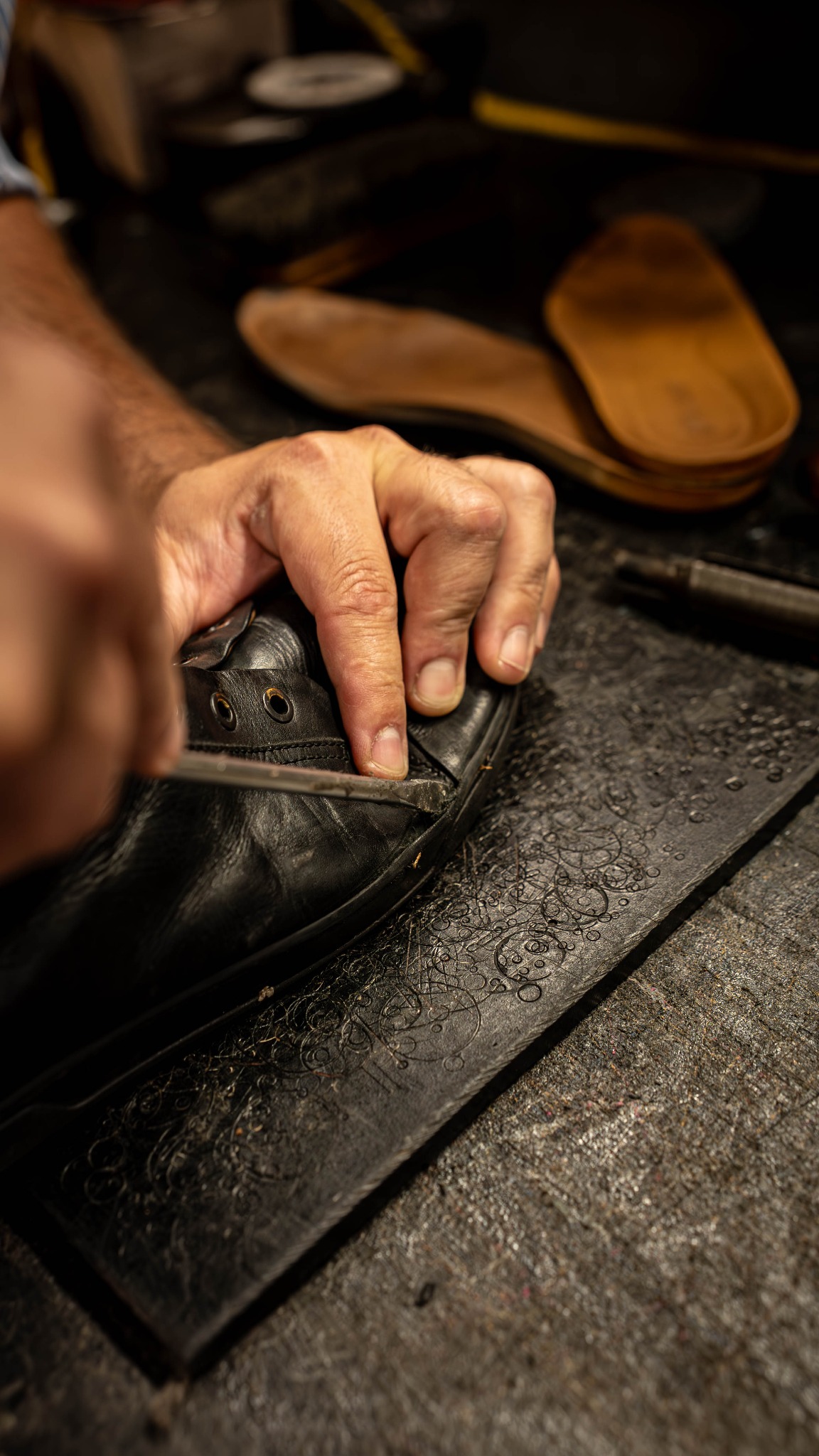 Czyszczenie czarnego skórzanego buta narzędziem na zdobionej płycie. W tle widoczne wkładki do butów. Detal pracy rzemieślniczej.