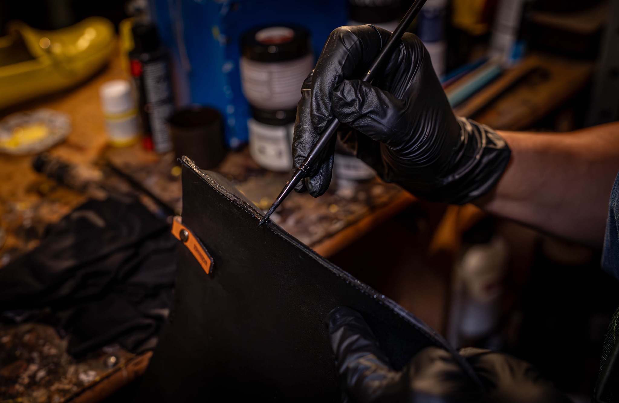 Ręczna praca nad skórzanym wyrobem: precyzyjne malowanie krawędzi czarną farbą za pomocą cienkiego pędzla, w tle warsztat z narzędziami i farbami.