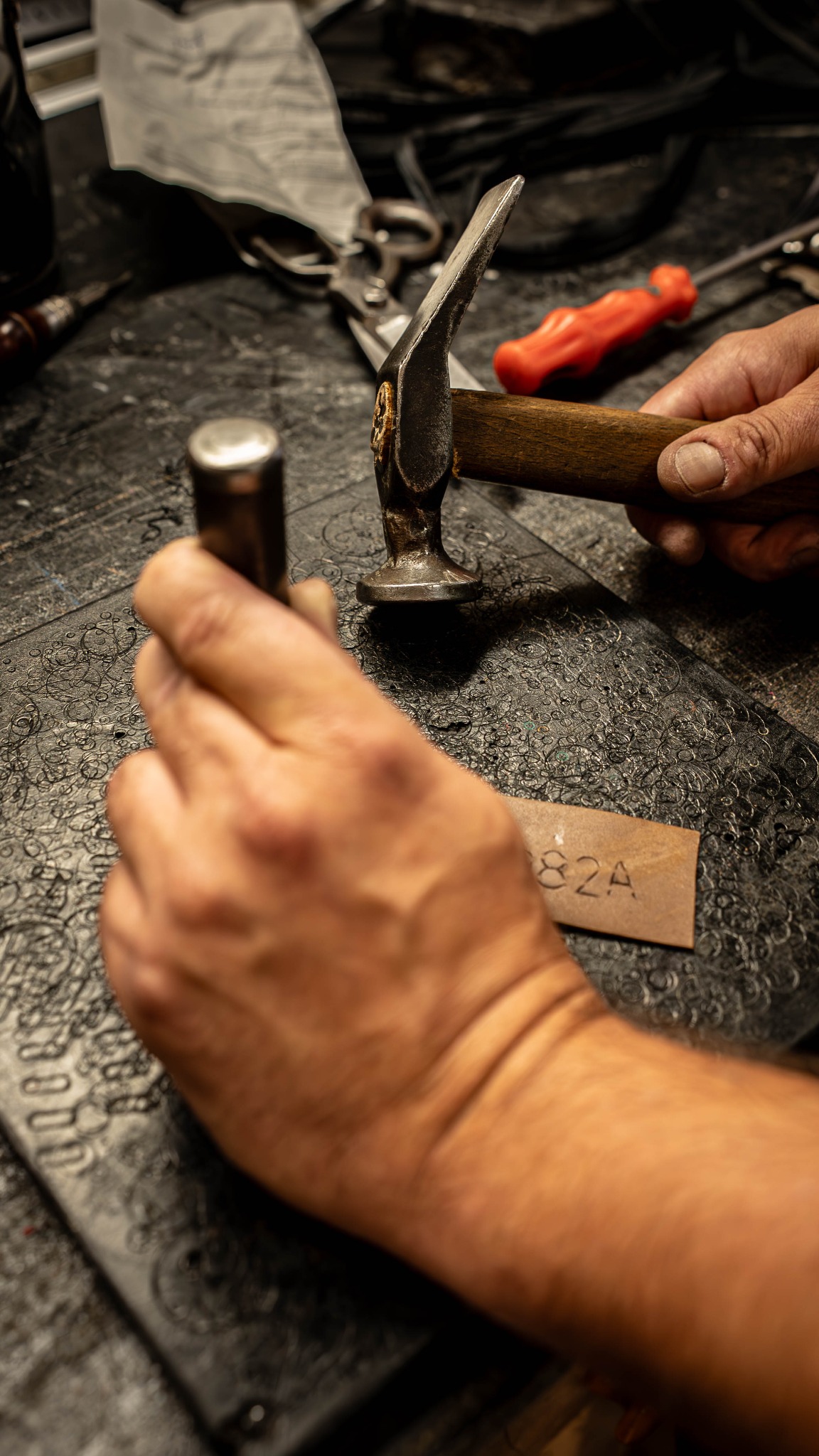 Ręczne zdobienie skóry narzędziami: młotek, stempel. Na stole roboczym skórzany fragment z numerem '82A'. Detal pracy rzemieślniczej.