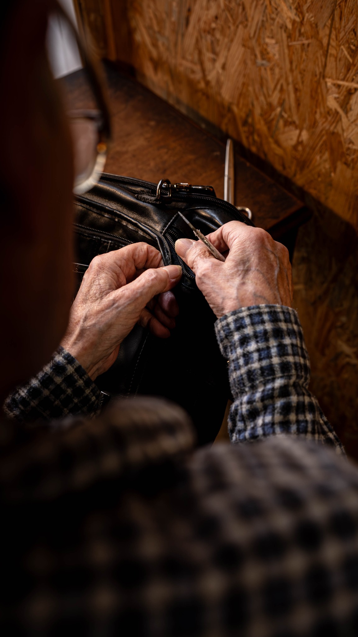 Ręce starszej osoby naprawiającej zamek błyskawiczny w czarnej skórzanej torbie na tle drewnianej ściany. Widoczne narzędzie i fragment ubrania w kratę.