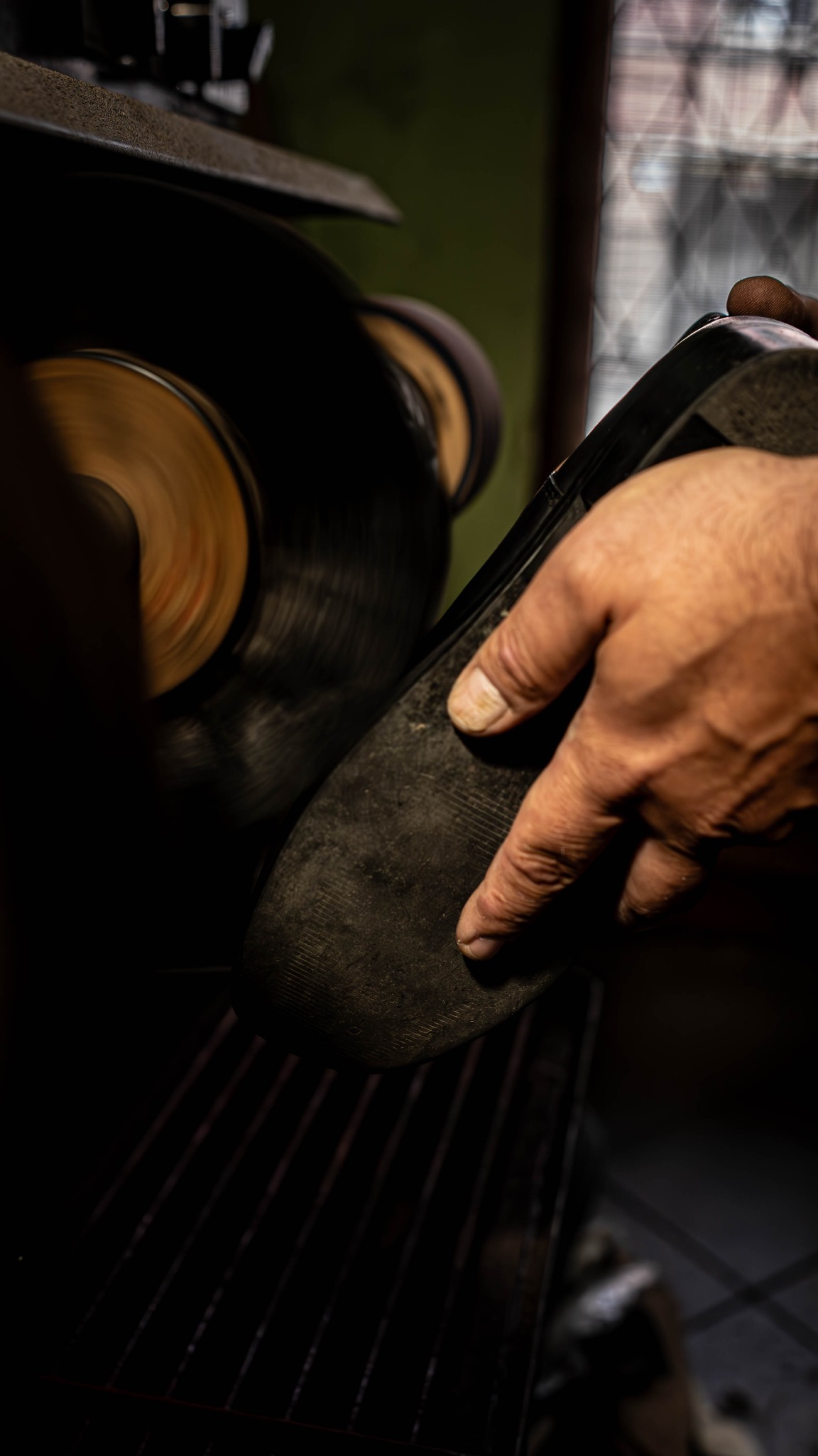 Polerowanie podeszwy buta na obracającej się szczotce przez rzemieślnika. Detal dłoni trzymającej but, w tle warsztat i okno.