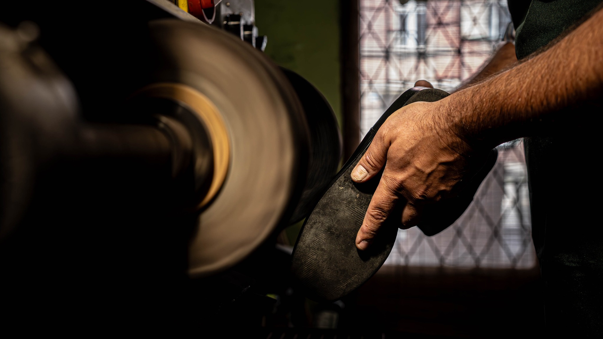 Ręka przytrzymuje podeszwę buta przy obracającej się polerce, widoczne detale skóry i gumy, w tle okno z kratą.