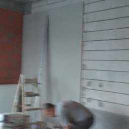 Marek - Montaż płyty G-K: ściana z płytą, drabina, osoba w szarej koszulce w trakcie pracy, widoczny fragment grzejnika i ceglanej ściany w surowym wnętrzu.