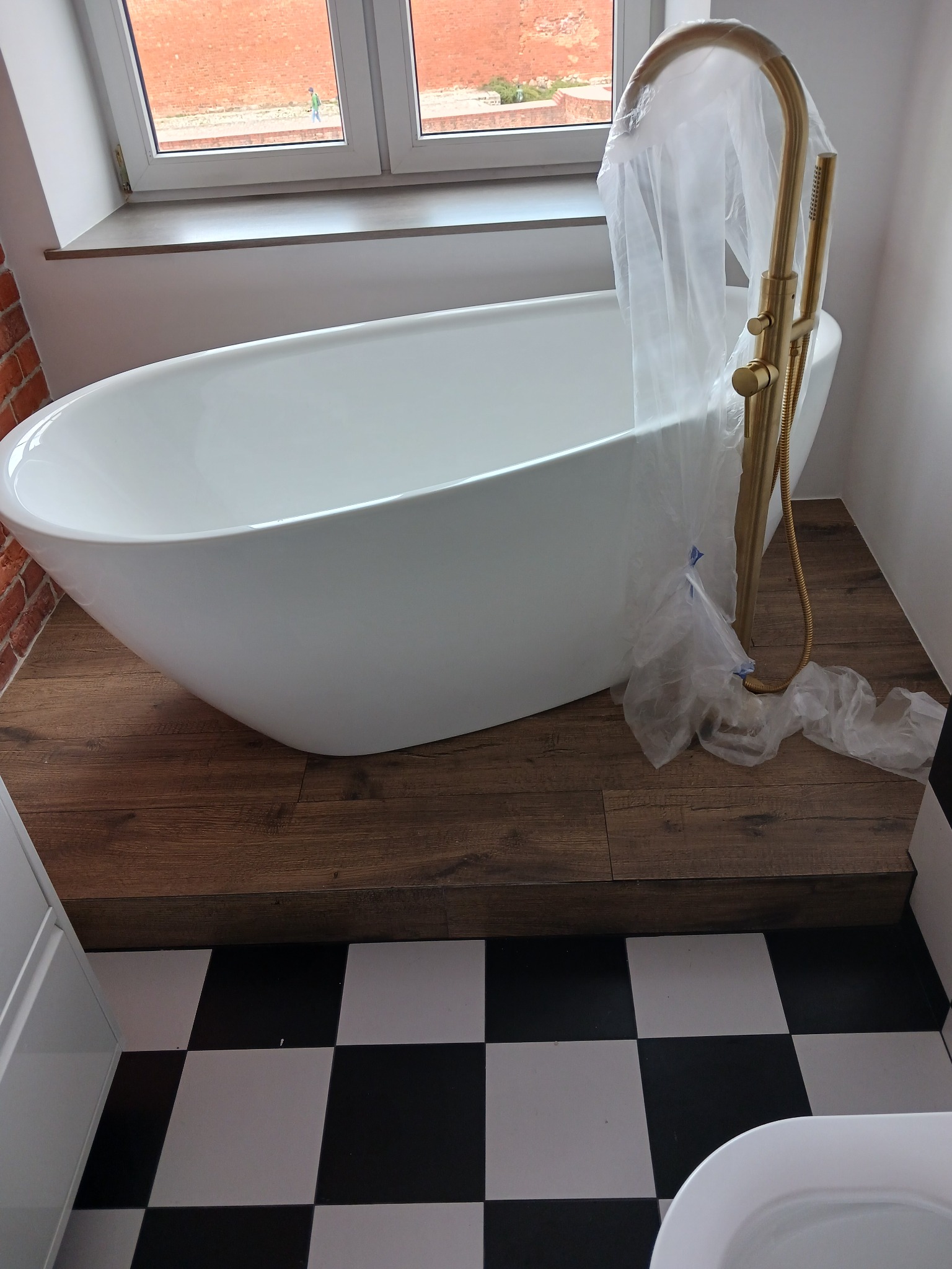 Nowoczesna, biała wanna wolnostojąca z złotą armaturą, zabezpieczona folią, na tle drewnopodobnej podłogi, z widoczną szachownicą płytek i ceglaną ścianą.