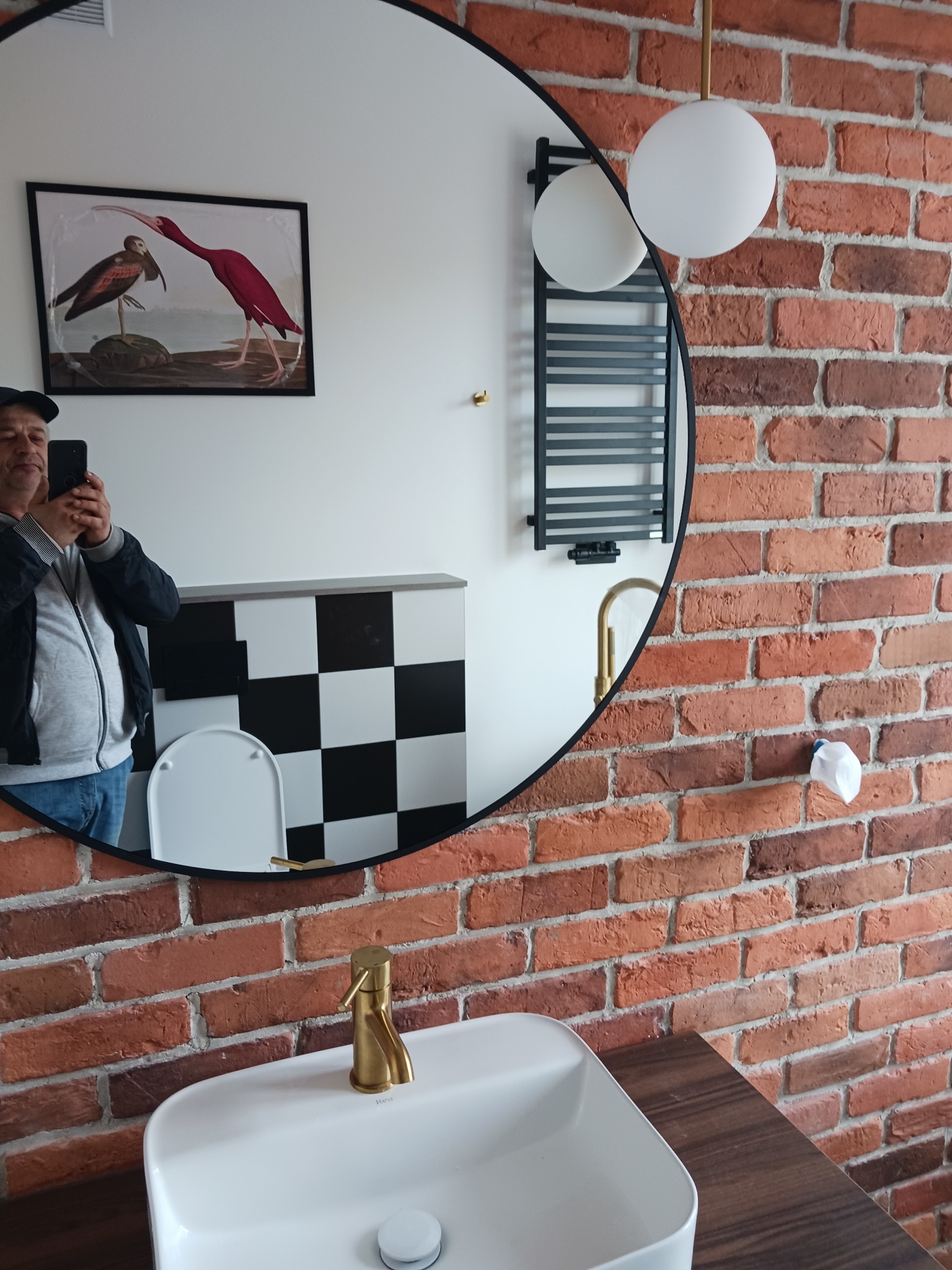 Okrągłe lustro z odbiciem mężczyzny robiącego zdjęcie w łazience z ceglaną ścianą, białą umywalką, złotą baterią i grzejnikiem drabinkowym.