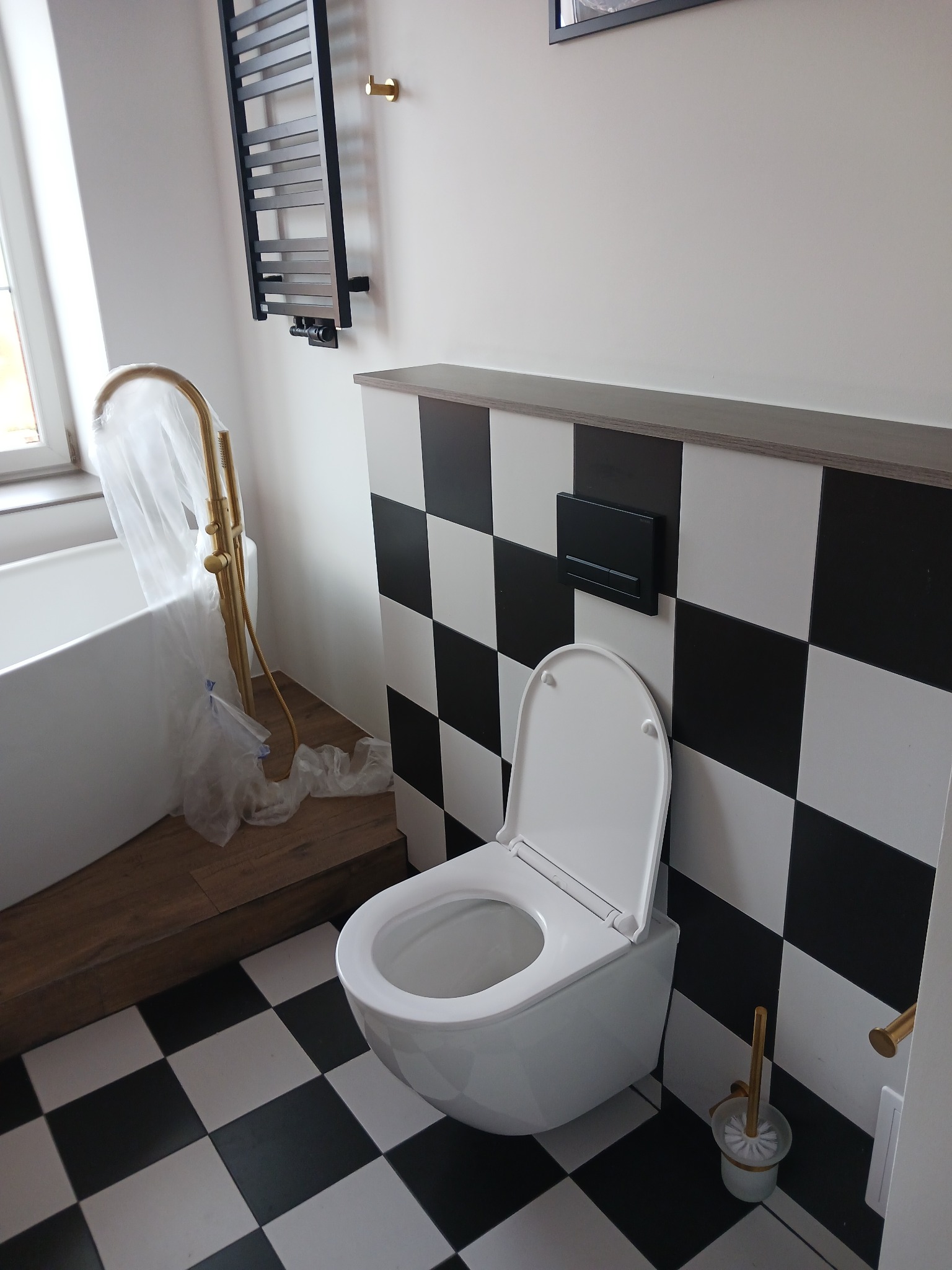 Nowoczesna łazienka z białą toaletą i czarno-białą szachownicą na podłodze i ścianie. Złota armatura częściowo osłonięta folią ochronną. Grzejnik drabinkowy na ścianie.