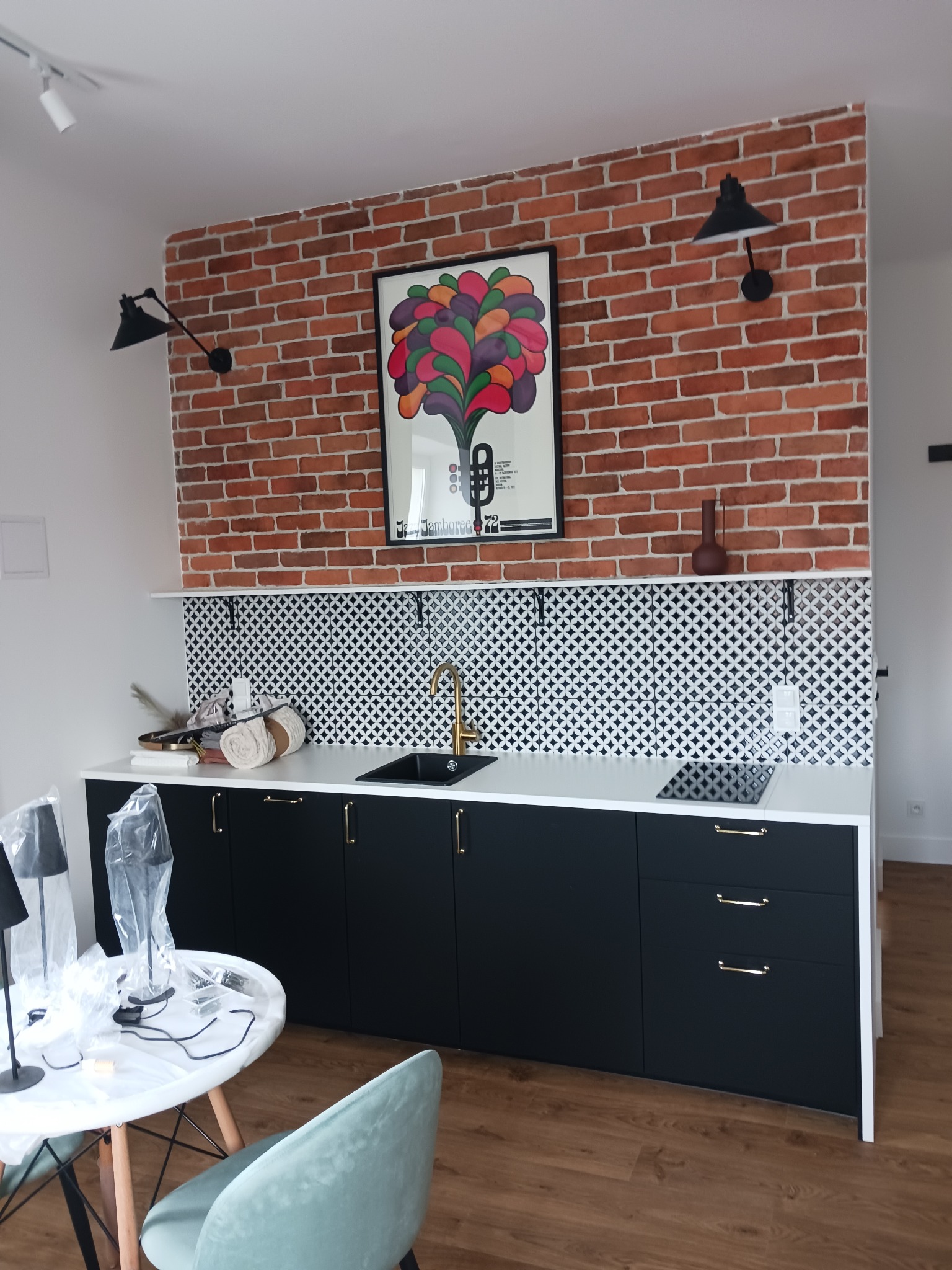 Nowoczesna kuchnia z ceglaną ścianą, czarnymi szafkami i złotą armaturą. Nad blatem biało-czarne płytki. Na ścianie plakat i lampy. Widoczne elementy wykończenia.