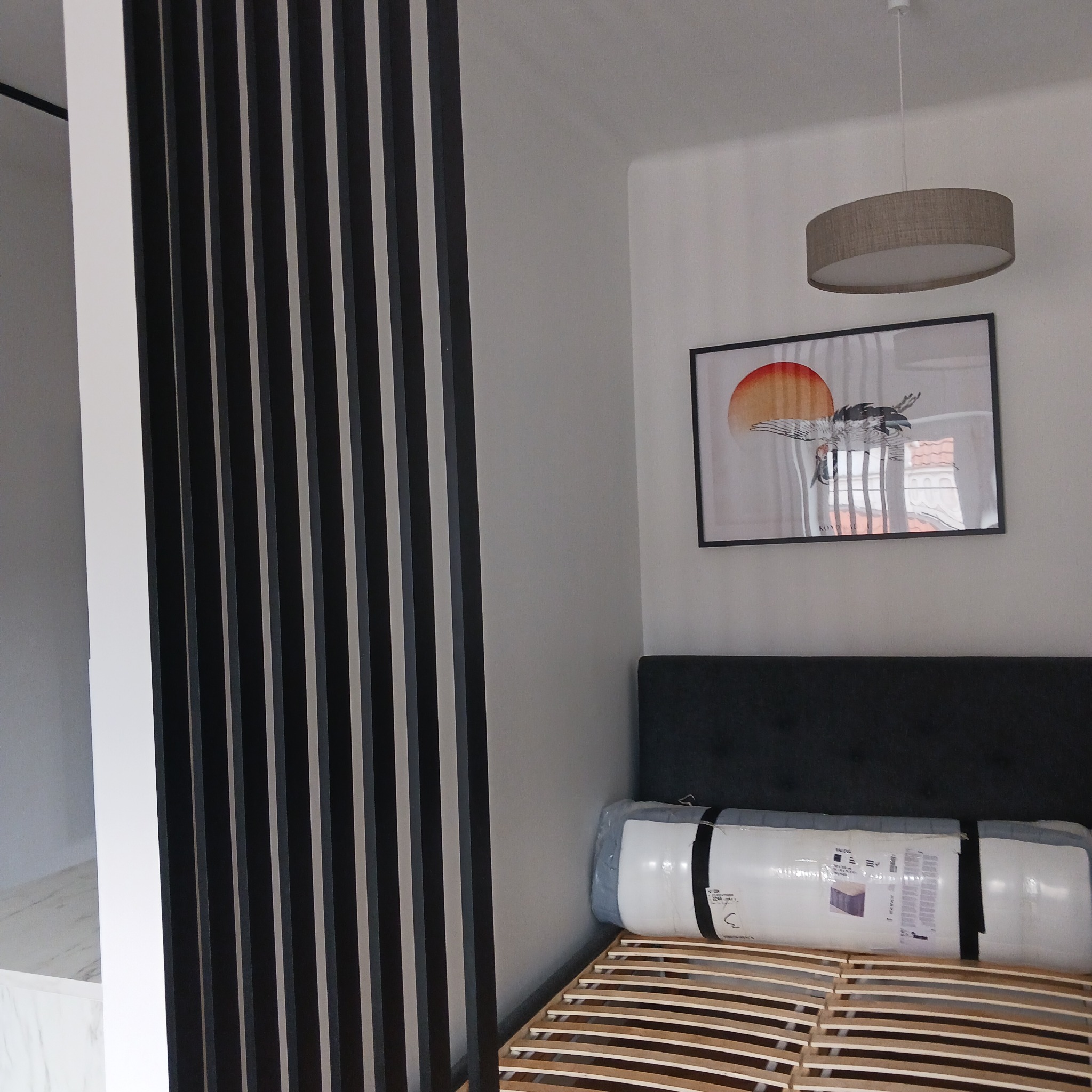 Nowoczesny pokój z drewnianą ażurową ścianką działową, łóżkiem ze stelażem i obrazem z motywem żurawia. Minimalistyczna lampa sufitowa. Jasne wnętrze.