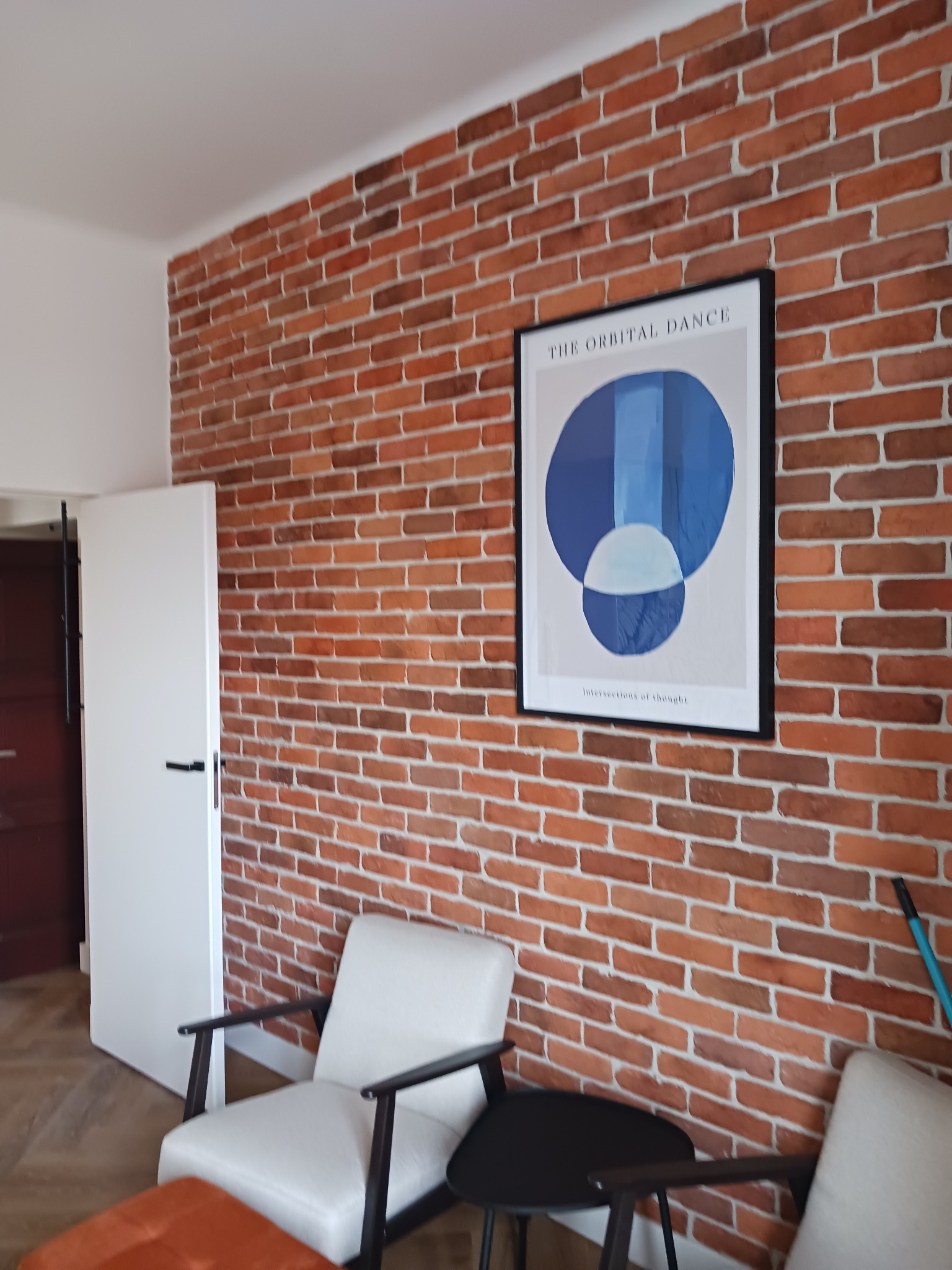 Ceglana ściana z dekoracyjnym wzorem, uzupełniona nowoczesnym plakatem i minimalistycznymi fotelami, tworzy ciepłe i stylowe wnętrze.