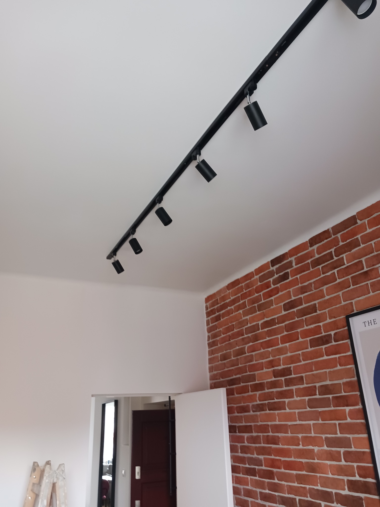 Czarna szyna z reflektorami sufitowymi, oświetlająca ścianę z cegły w jasnym wnętrzu. Widoczny fragment białej ściany i drzwi w tle.