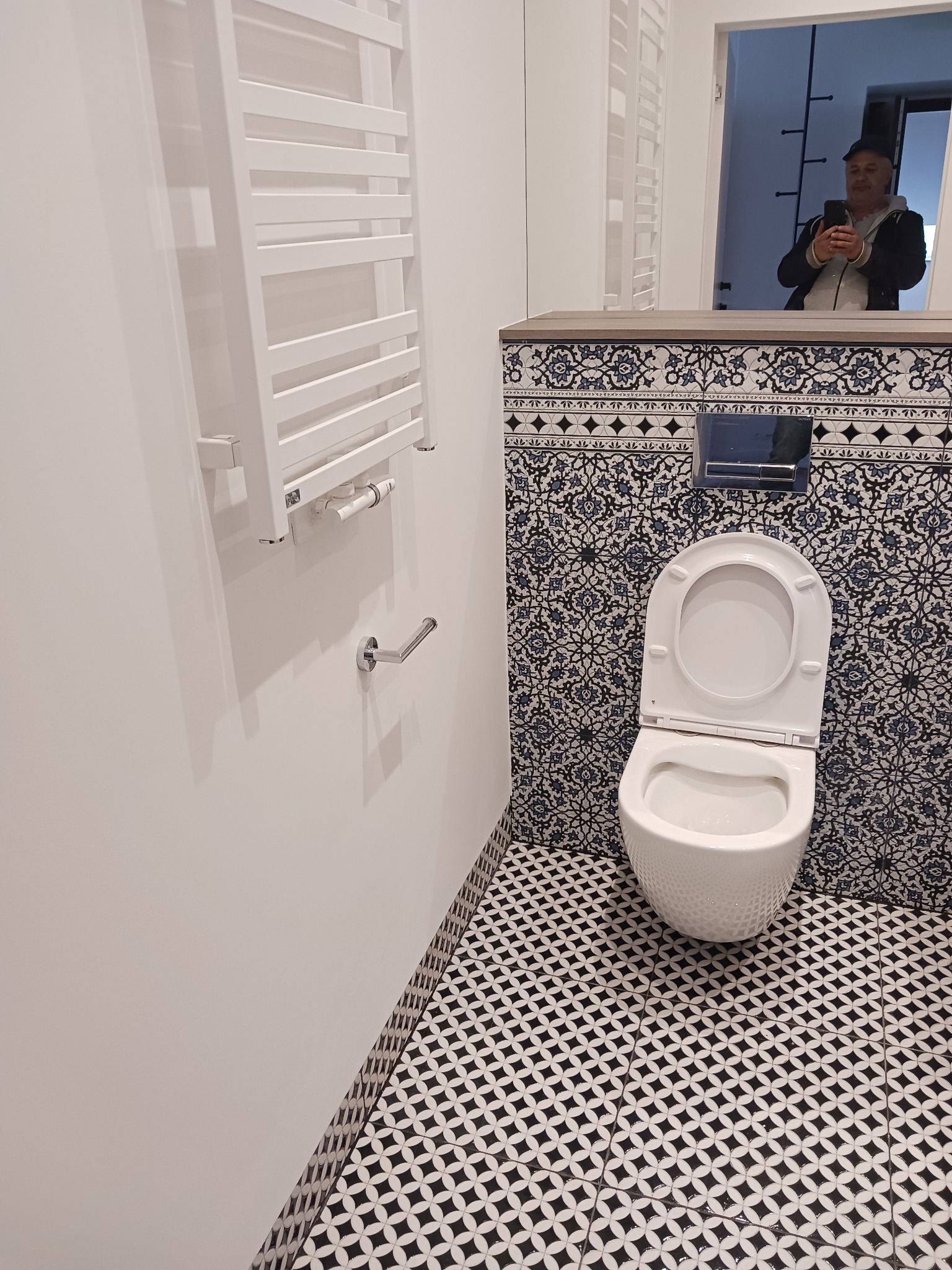 Biała toaleta z deską w górze, wzorzyste płytki na ścianie i podłodze w odnowionej łazience. W lustrze odbija się osoba trzymająca telefon.