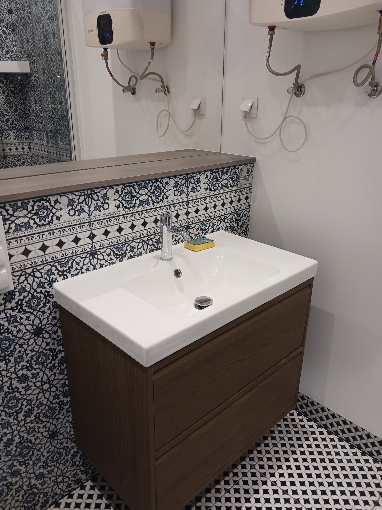 Nowoczesna łazienka po remoncie: biała umywalka z drewnianą szafką, dekoracyjne płytki na ścianie i podłodze, bojler elektryczny na ścianie.