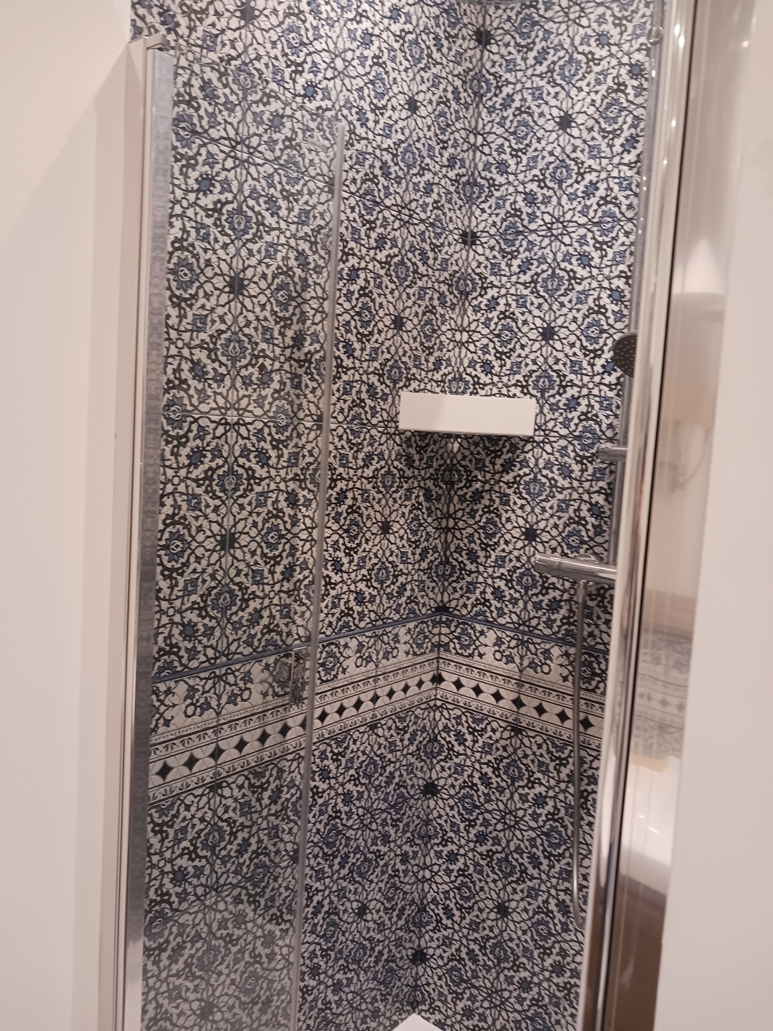 Wykończona kabina prysznicowa z wzorzystą, niebiesko-białą glazurą w Ząbkach. Widoczna półka i chromowana armatura. Nowoczesny design i dbałość o detale.