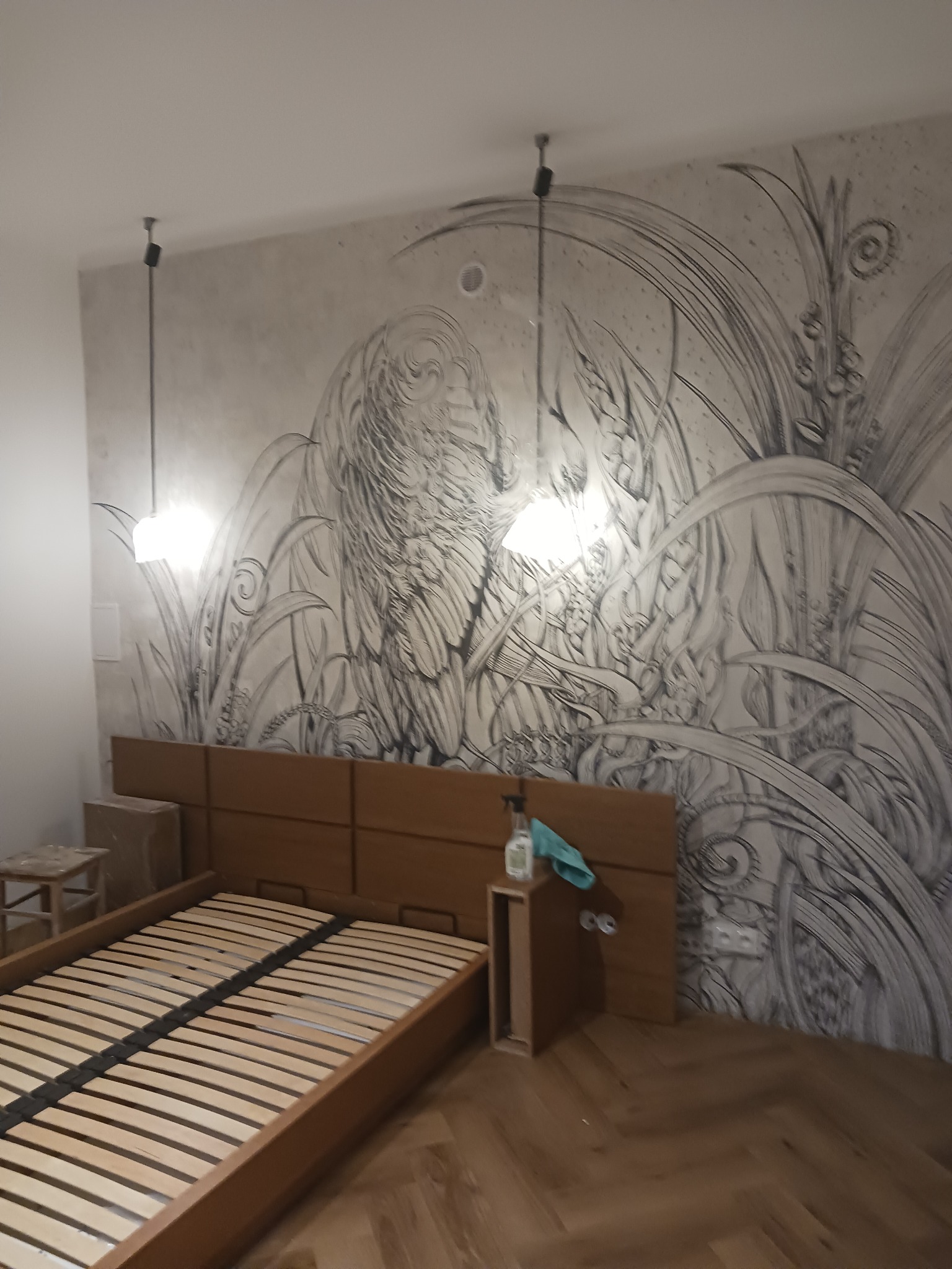 Sypialnia z drewnianym łóżkiem i oryginalną, ręcznie malowaną grafiką na ścianie. Parkiet ułożony w jodełkę, lampy wiszące. Widoczne akcesoria do sprzątania.