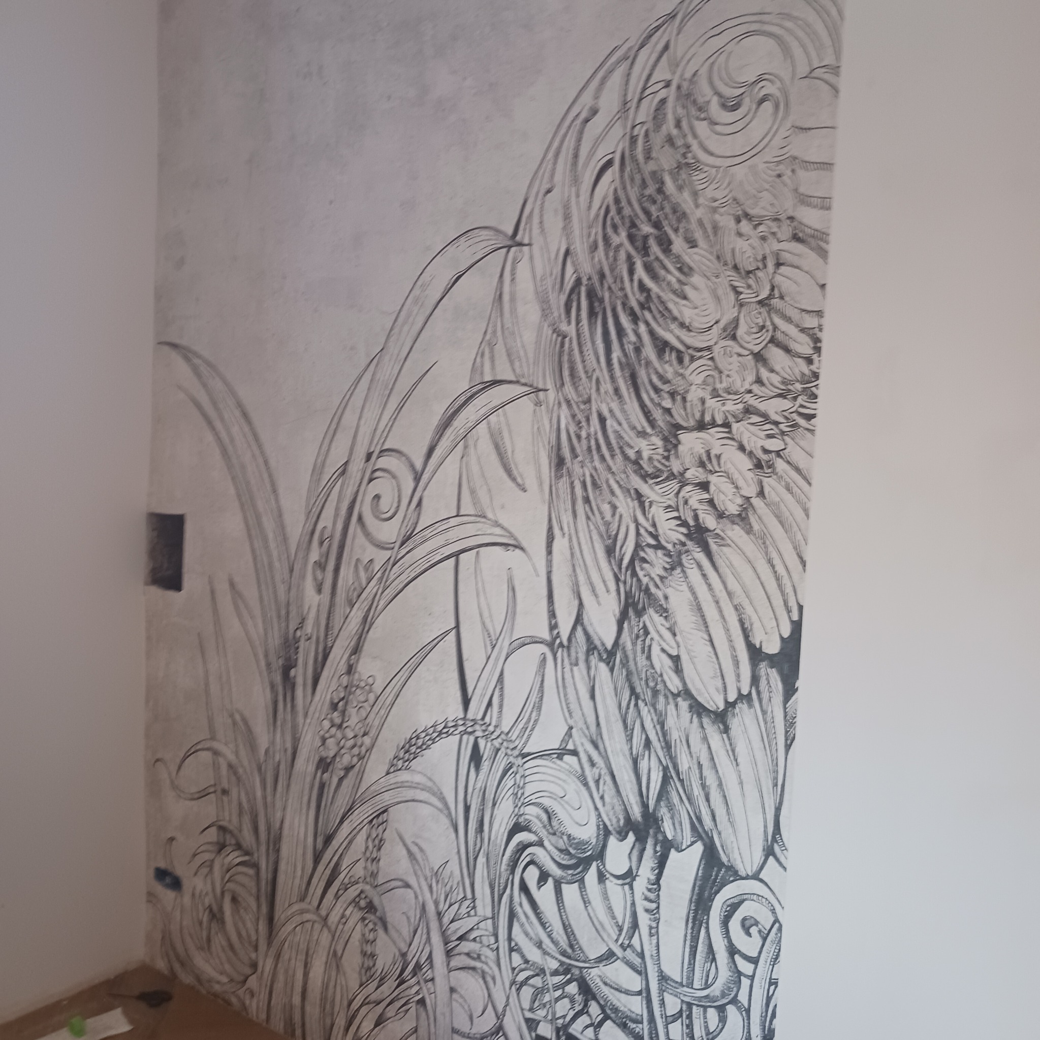 Ściana z fototapetą w odcieniach szarości, przedstawiającą rysunek żurawia w trawie. Widoczny fragment gniazdka elektrycznego i podłoga w rogu.