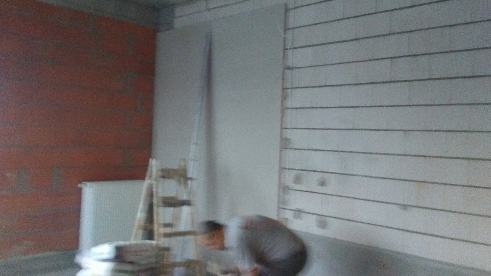 Montaż płyty G-K: ściana z płytą, drabina, osoba w szarej koszulce w trakcie pracy, widoczny fragment grzejnika i ceglanej ściany w surowym wnętrzu.