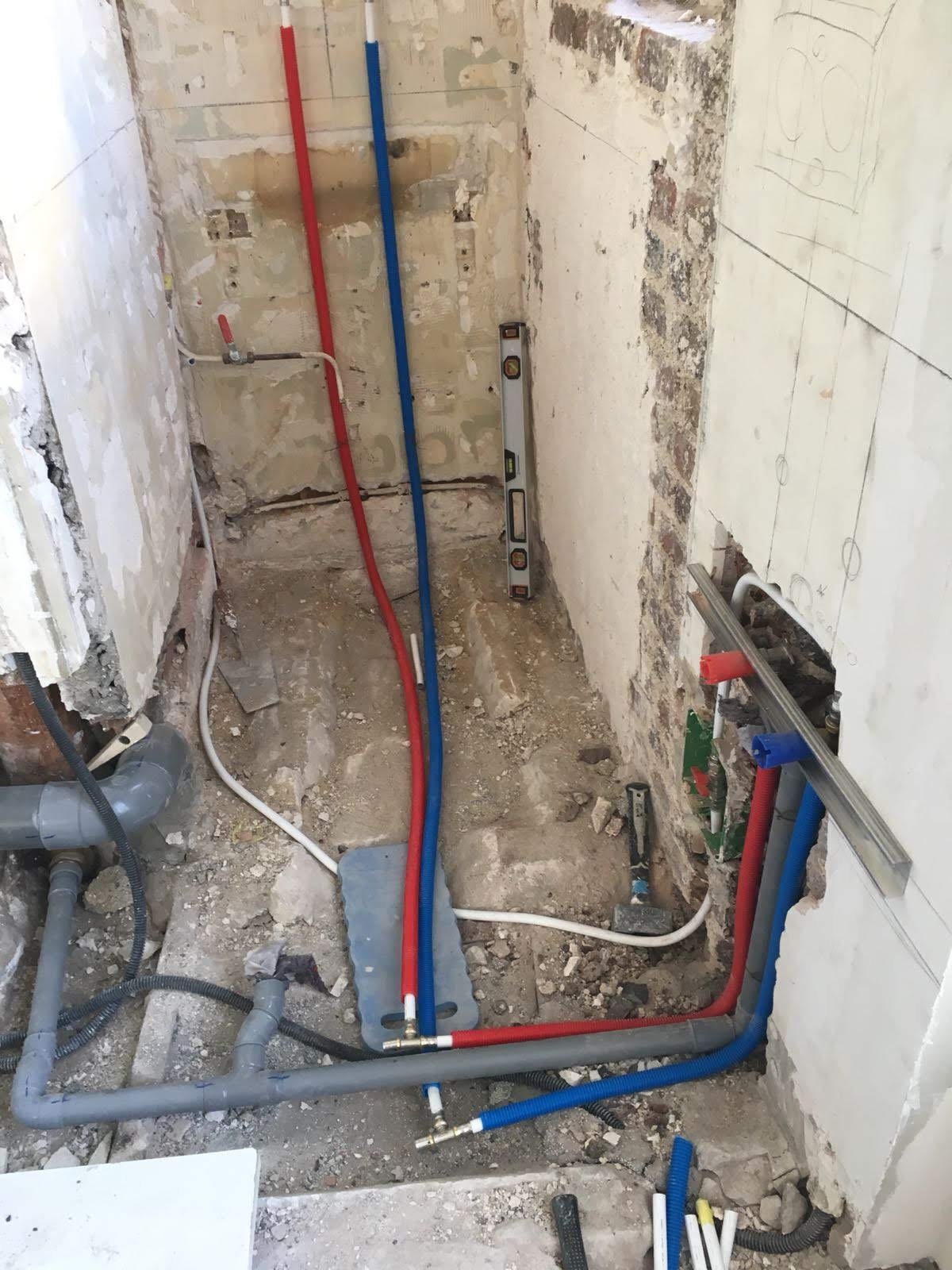 Wnętrze w trakcie prac: czerwone i niebieskie rury instalacji wodnej prowadzone wzdłuż surowych ścian. Widoczny poziomica i fragment instalacji kanalizacyjnej.