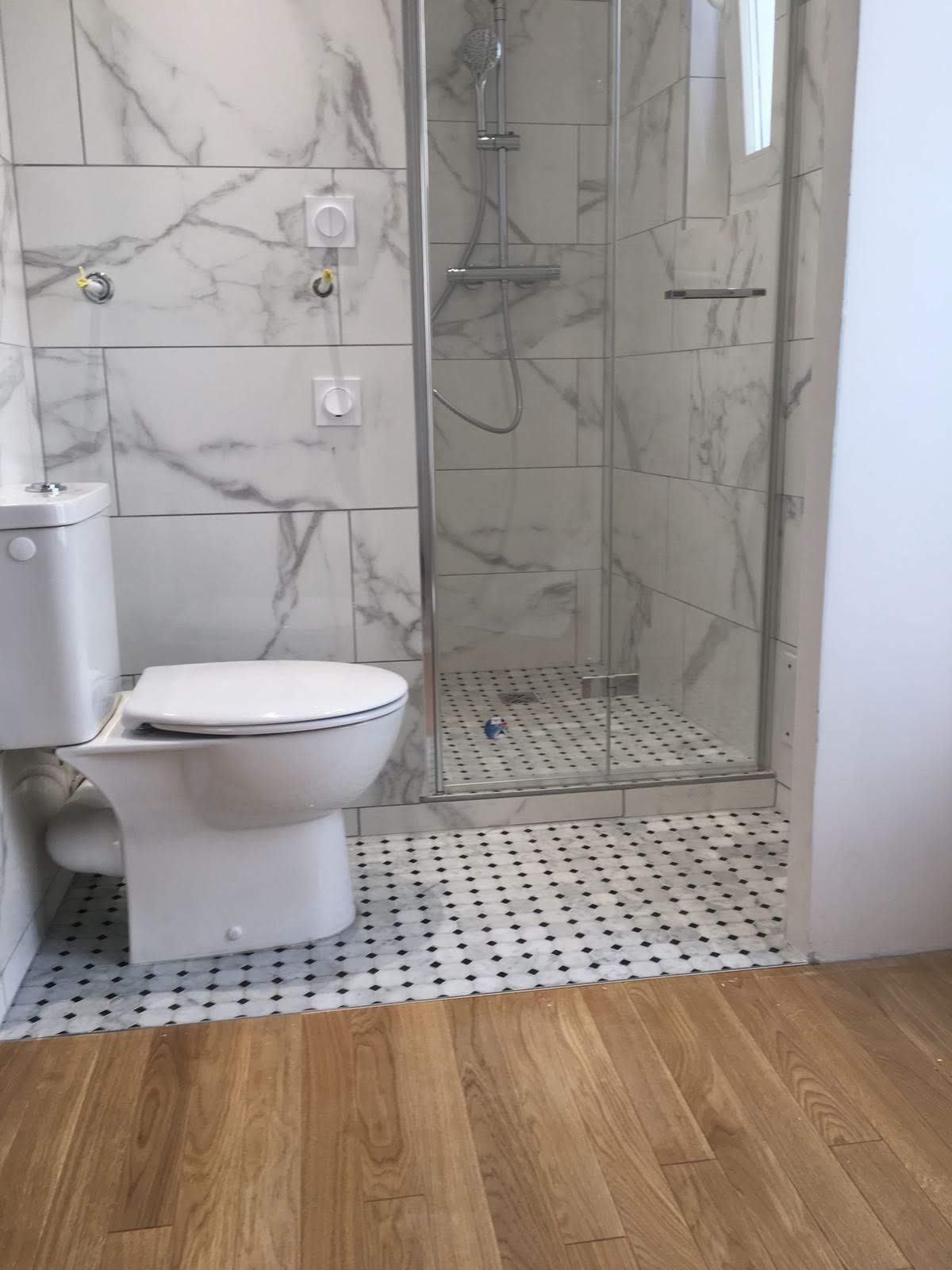 Biała toaleta i kabina prysznicowa z marmurowymi płytkami ściennymi oraz podłogą w czarno-białą mozaikę. Panele podłogowe w odcieniach brązu. Widok na fragment łazienki.