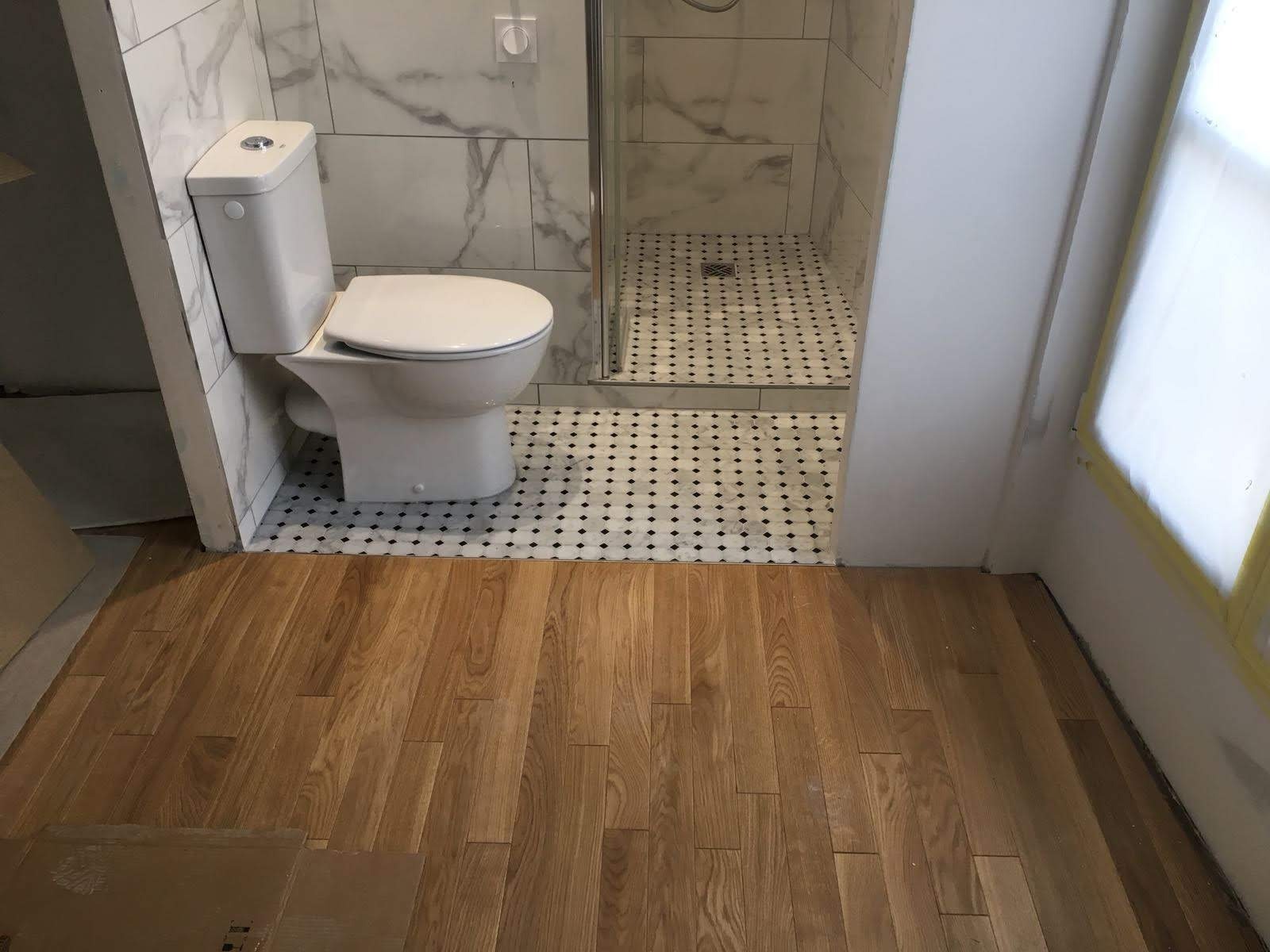 Nowoczesna łazienka po remoncie: marmurowe płytki, kabina prysznicowa z mozaiką, drewniana podłoga. Widok na toaletę i fragment okna z taśmą malarską.