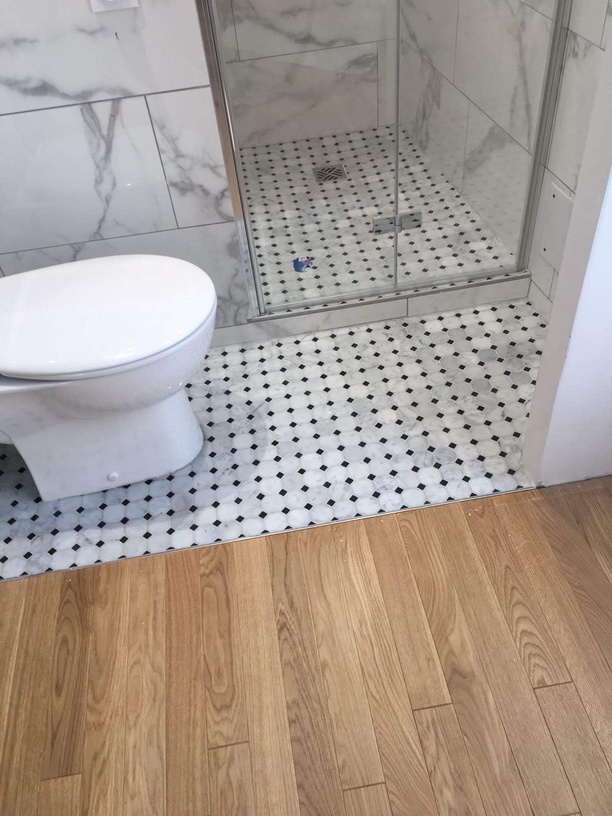 Biała toaleta sąsiaduje z kabiną prysznicową wyłożoną marmurowymi płytkami i mozaiką. Podłoga z paneli drewnianych. Widok z góry.