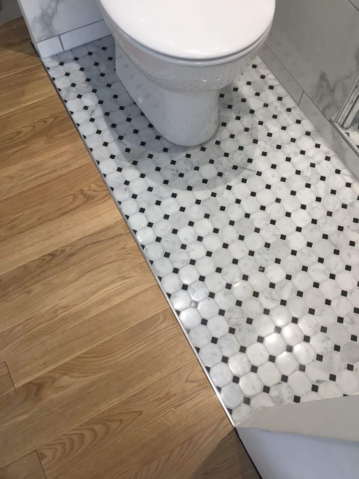 Biało-czarna mozaika heksagonalna na podłodze w łazience, obok drewniane panele, oddzielone metalową listwą. Widoczny fragment toalety i marmurowe wykończenie ściany.