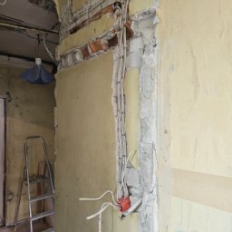 IVAN KRASNOSHCHOKOV - Ściana w trakcie remontu: odsłonięta cegła, instalacja elektryczna w peszlach, fragment ściany z resztkami starej farby. Widoczna drabina i wisząca lampa.