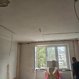 IVAN KRASNOSHCHOKOV - Pokój w trakcie remontu: ściany przygotowane do malowania, widoczne przewody elektryczne, okno z widokiem na zieleń, narzędzia i materiały budowlane.