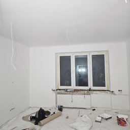 IVAN KRASNOSHCHOKOV - Wnętrze po malowaniu na biało. Widoczne zaklejone okno, grzejnik owinięty folią, narzędzia malarskie i worki z zaprawą na podłodze. Kabel zwisa z sufitu.