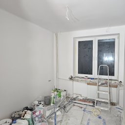 IVAN KRASNOSHCHOKOV - Pokój po malowaniu, z widocznymi śladami prac: zakryte okno, drabina, farby. Świeżo pomalowane ściany na biało. Etap wykończeniowy przed sprzątaniem.