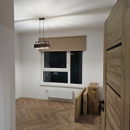 IVAN KRASNOSHCHOKOV - Pokój po remoncie: parkiet w jodełkę, białe ściany, okno z roletą, nowoczesna lampa. Częściowo widoczne drzwi i kartony z wyposażeniem.